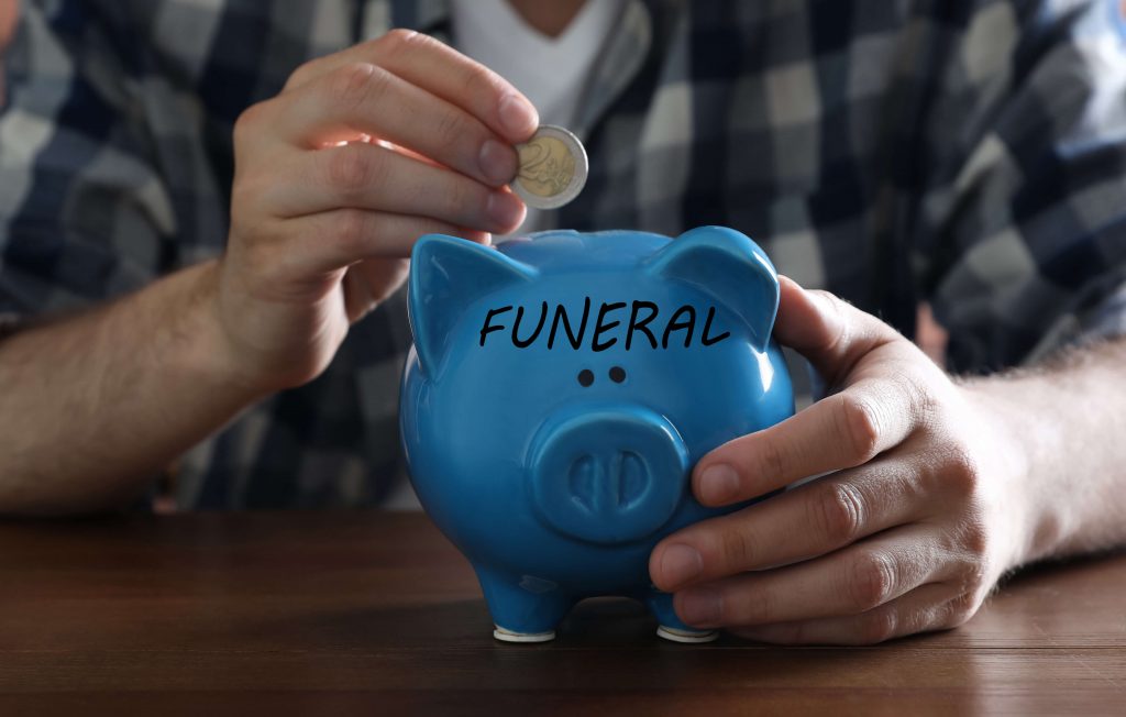 Person wirft eine Münze in eine Spardose mit der Aufschrift Funeral für Bestattungskosten

